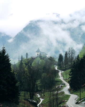 A serene mountain landscape with a winding path leading through lush, green trees and a small stream flowing alongside. In the distance, a quaint building with a pointed roof sits atop a hill. Mist and clouds hover over the scene, partially obscuring the mountain peaks and adding an ethereal feel.