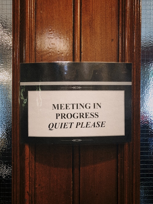 A sign is attached to a wooden door, requesting silence as a meeting is in progress. The text is printed in black capital letters on a white background, bordered with a simple black design.