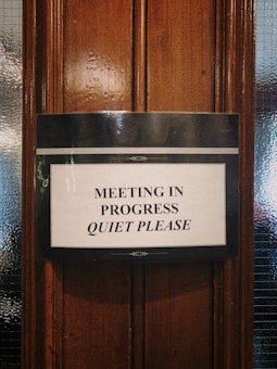 A sign is attached to a wooden door, requesting silence as a meeting is in progress. The text is printed in black capital letters on a white background, bordered with a simple black design.