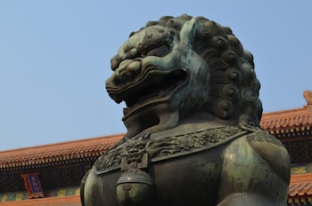 A large, intricately detailed bronze lion statue with a fierce expression is positioned in front of an ornate, historical building with traditional Asian architecture. The building features a red tiled roof and a sign with Chinese characters, set against a clear blue sky.