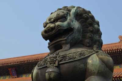 A large, intricately detailed bronze lion statue with a fierce expression is positioned in front of an ornate, historical building with traditional Asian architecture. The building features a red tiled roof and a sign with Chinese characters, set against a clear blue sky.