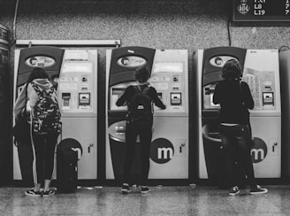 Three people are standing in front of ticket vending machines, interacting with the screens. The scene is set in a subway or train station with a textured wall background. Each person is wearing casual attire with backpacks, and there is luggage placed next to them.