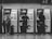 Three people are standing in front of ticket vending machines, interacting with the screens. The scene is set in a subway or train station with a textured wall background. Each person is wearing casual attire with backpacks, and there is luggage placed next to them.