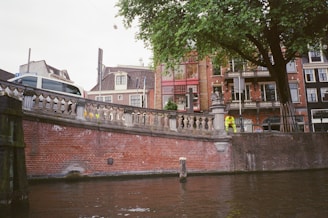 A scenic urban canal scene with a red brick wall lining the waterway. Above the wall, a historic cityscape with a mixture of traditional architecture, including red brick and timber-framed buildings. A person in high-visibility clothing stands near a stone balustrade. Leafy green trees add a natural element to the streetscape.
