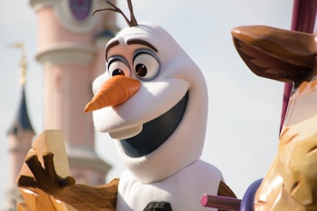A cheerful snowman character with a carrot nose and large eyes is part of a parade or display. The background features a tower reminiscent of castle architecture, suggesting a theme park setting.