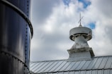 A close-up view of a metal rooftop with an old weathered ventilation structure on top, featuring a small antenna or lightning rod. The structure has noticeable rust and paint peeling. To the left, part of a large, dark cylindrical metal silo appears, with rivets along its seam. The sky is partly cloudy with some blue patches visible.