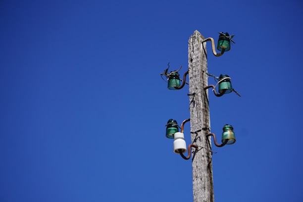 A tall wooden utility pole stands against a clear blue sky. The pole is equipped with several glass insulators and wires, which are arranged neatly in pairs along its length. The insulators are primarily green with one white insulator visible.