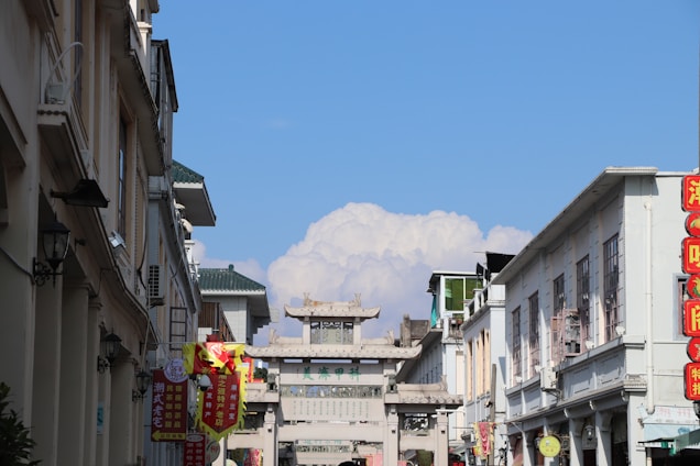 A bustling urban street scene with traditional architectural elements, including a prominent stone gateway adorned with intricate designs. Tall buildings line both sides, featuring a mix of modern and classic Chinese styles. Bright red and yellow banners hang along the street, written in Chinese characters. In the background, fluffy clouds fill the blue sky.