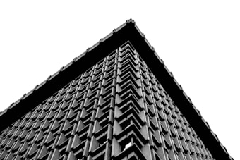 A black and white photograph of a building with a highly textured facade, featuring a repeating geometric pattern. The image focuses on the corner of the structure, emphasizing the angular lines and depth of the architecture against a stark white sky.