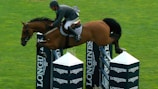 A horseback rider dressed in equestrian attire is leaping over a series of barriers labeled with 'LONGINES' in a grassy field. The horse is mid-air, demonstrating athleticism and agility as it clears the high jump with ease.