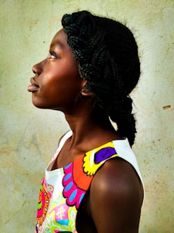 A person with braided hair is shown in profile against a textured, earthy wall. The subject is wearing a colorful, patterned garment with vibrant colors.