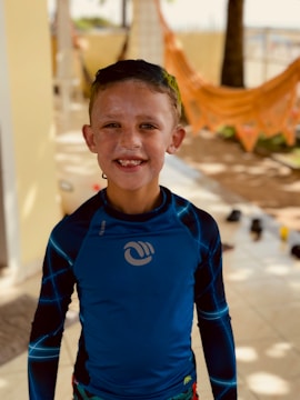 A young boy with a smiling expression stands indoors wearing a blue rash guard with stylish patterns on the sleeves. His face is covered with sunscreen, and his hair is wet. In the background, there's an orange hammock, some blurred furniture, and a sunny environment.