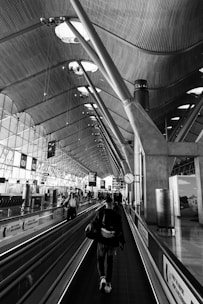 A spacious and modern airport terminal with architectural elements featuring curved ceilings, exposed beams, and a series of large windows allowing natural light to flood the area. A few travelers can be seen walking along a moving walkway and the floor area, alongside signage directing passengers to gates. The setting is busy with multiple lights and displays contributing to the futuristic ambiance.