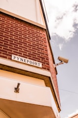 A corner of a brick building featuring a sign with the word 'PYNEPOORT'. A security camera is mounted on the wall, and part of the sky with clouds is visible.