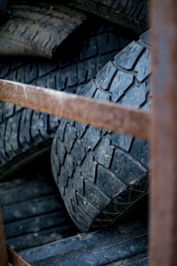 A collection of stacked, worn-out tires with visible tread patterns. The tires appear old and used, with a rugged texture. There is a rusty metal frame partially obscuring the view in the foreground.