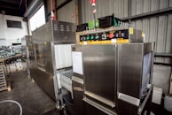 A large industrial machine with a metallic finish, featuring control panels with buttons, switches, and temperature displays. The setting appears to be a warehouse or factory space with metal walls and shelving.