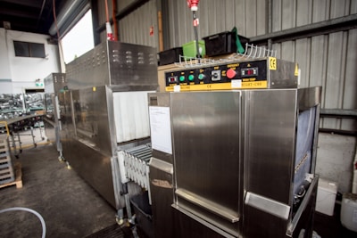 A large industrial machine with a metallic finish, featuring control panels with buttons, switches, and temperature displays. The setting appears to be a warehouse or factory space with metal walls and shelving.