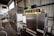 A large industrial machine with a metallic finish, featuring control panels with buttons, switches, and temperature displays. The setting appears to be a warehouse or factory space with metal walls and shelving.