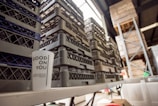 A white cup with the text 'GOOD ON YOU' is placed on a table in a warehouse setting. Several stacks of gray and black plastic crates are arranged behind the cup. Part of a metal shelving unit and some wooden pallets are visible in the background, suggesting an industrial or storage environment.