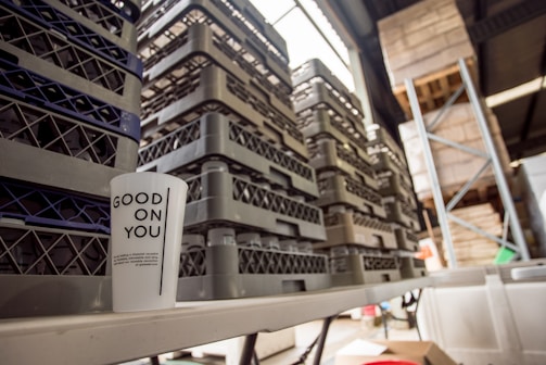 A white cup with the text 'GOOD ON YOU' is placed on a table in a warehouse setting. Several stacks of gray and black plastic crates are arranged behind the cup. Part of a metal shelving unit and some wooden pallets are visible in the background, suggesting an industrial or storage environment.