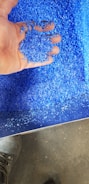 A hand holding a pile of bright blue plastic granules over a larger collection of the same material. The granules have a consistent size and texture, suggesting they are industrially processed. The background shows more of the granules scattered around, with a small portion of a shoe visible on a gray floor.