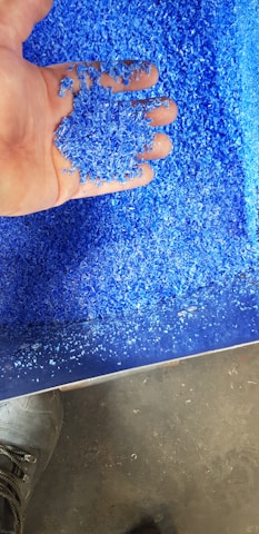 A hand holding a pile of bright blue plastic granules over a larger collection of the same material. The granules have a consistent size and texture, suggesting they are industrially processed. The background shows more of the granules scattered around, with a small portion of a shoe visible on a gray floor.