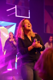 Kelly Slusher mid-performance, microphone in hand, with a joyful expression under warm stage lights.