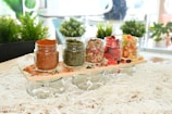 Five small glass jars on a wooden tray hold various colorful dried spices and fruits. They are placed on a tabletop with a background of lush green plants and a bright, airy interior. The contents of the jars range from red and green spices to multicolored dried fruits.