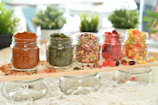 Assortment of homemade condiments in glass jars with fresh herbs and fruits around.