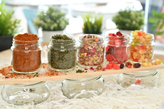 Five small glass jars are arranged on a wooden plank, each filled with different colorful ingredients, including red chili powder, green herbs, mixed dried flowers, red and black berries, and an assortment of dried fruits. The background features blurred greenery, providing a fresh and natural setting.