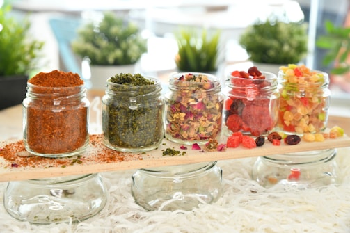 Five small glass jars are arranged on a wooden plank, each filled with different colorful ingredients, including red chili powder, green herbs, mixed dried flowers, red and black berries, and an assortment of dried fruits. The background features blurred greenery, providing a fresh and natural setting.