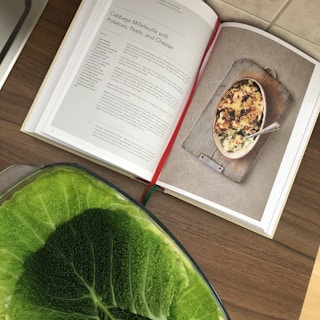 An open cookbook displays a recipe for 'Cabbage Millefeuille with Potatoes, Pesto, and Cheese.' The image on the right page shows the completed dish, presented in a rustic baking dish. Below the book, a large green cabbage is partially visible, adding a fresh element to the scene.