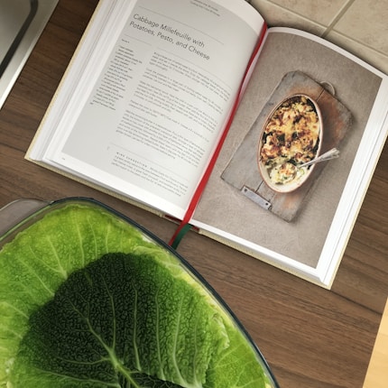 An open cookbook displays a recipe for 'Cabbage Millefeuille with Potatoes, Pesto, and Cheese.' The image on the right page shows the completed dish, presented in a rustic baking dish. Below the book, a large green cabbage is partially visible, adding a fresh element to the scene.