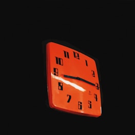 A red square wall clock with bold black numbers and hands is set against a dark background. The number design appears vintage, and the clock gives off a retro aesthetic. Bright highlights on the clock's surface suggest a glossy finish.