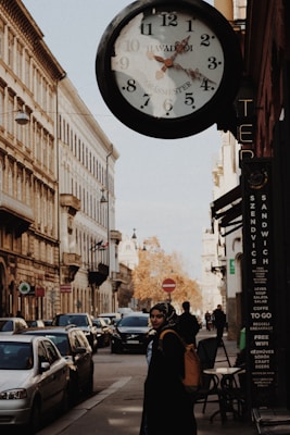 A street scene with historic buildings lining both sides. A large vintage clock is prominently hanging above a sidewalk, showing the time close to 2:22. The street is busy with parked cars and a few moving vehicles. A person carrying a backpack is walking on the sidewalk, while another person is visible in the background. There is a signboard on the side of a building advertising sandwiches and craft beers.