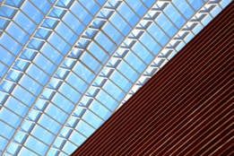A glass-paneled ceiling with a geometric pattern of rectangular glass sections supported by metal frames. Below the glass ceiling is a slanted wooden structure featuring horizontal slats.