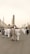 A large group of people dressed in white traditional garments walk towards an ornate architectural structure. The scene includes several tall minarets in the background under a clear, light-colored sky, suggesting a religious or cultural gathering.