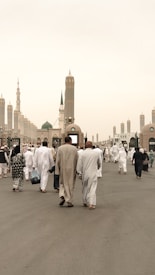 A large group of people dressed in white traditional garments walk towards an ornate architectural structure. The scene includes several tall minarets in the background under a clear, light-colored sky, suggesting a religious or cultural gathering.