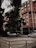 A red brick residential building with multiple floors is surrounded by lush trees. Several cars are parked in the foreground in a concrete driveway, while the branches of the trees obscure part of the structure.
