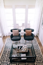 Two modern armchairs with brown patterned cushions are positioned symmetrically on a geometric patterned rug. A glass coffee table holding various decorative items is placed in front of the chairs. The background features large windows with sheer white curtains allowing natural light to illuminate the room.