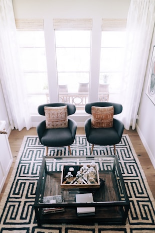 Two modern armchairs with brown patterned cushions are positioned symmetrically on a geometric patterned rug. A glass coffee table holding various decorative items is placed in front of the chairs. The background features large windows with sheer white curtains allowing natural light to illuminate the room.