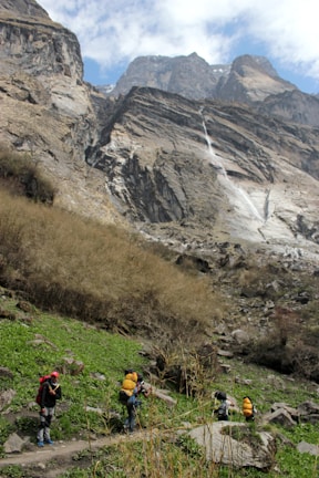 A group of hikers with backpacks is trekking through a mountainous landscape. The scene features tall, rugged cliffs and a cascading waterfall in the background, while the foreground shows green grass and rocky terrain. The sky is partially cloudy.