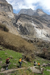A group of hikers with backpacks is trekking through a mountainous landscape. The scene features tall, rugged cliffs and a cascading waterfall in the background, while the foreground shows green grass and rocky terrain. The sky is partially cloudy.
