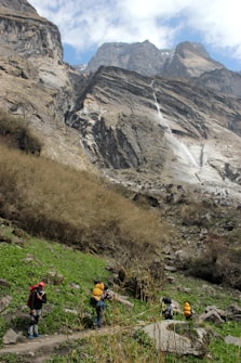A group of hikers with backpacks is trekking through a mountainous landscape. The scene features tall, rugged cliffs and a cascading waterfall in the background, while the foreground shows green grass and rocky terrain. The sky is partially cloudy.