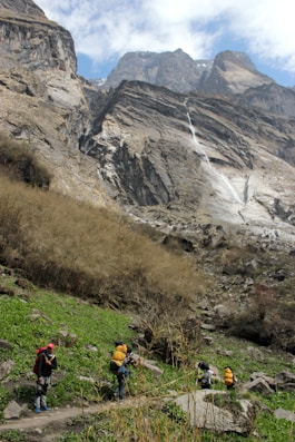 A group of hikers with backpacks is trekking through a mountainous landscape. The scene features tall, rugged cliffs and a cascading waterfall in the background, while the foreground shows green grass and rocky terrain. The sky is partially cloudy.