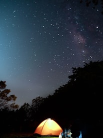 A glowing orange tent is set up under a vast, star-filled night sky. The stars are bright and numerous, casting a serene and tranquil atmosphere. Silhouettes of trees frame the scene, adding depth and context to the outdoor setting.