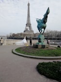A couple in wedding attire poses in front of the Eiffel Tower. The bride is wearing a white gown with a long train. A photographer is capturing the moment, and the scene includes a statue of a winged horse nearby. Trees and a cloudy sky form the backdrop.