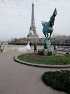 A couple in wedding attire poses in front of the Eiffel Tower. The bride is wearing a white gown with a long train. A photographer is capturing the moment, and the scene includes a statue of a winged horse nearby. Trees and a cloudy sky form the backdrop.