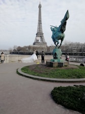A couple in wedding attire poses in front of the Eiffel Tower. The bride is wearing a white gown with a long train. A photographer is capturing the moment, and the scene includes a statue of a winged horse nearby. Trees and a cloudy sky form the backdrop.