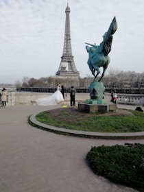 A couple in wedding attire poses in front of the Eiffel Tower. The bride is wearing a white gown with a long train. A photographer is capturing the moment, and the scene includes a statue of a winged horse nearby. Trees and a cloudy sky form the backdrop.